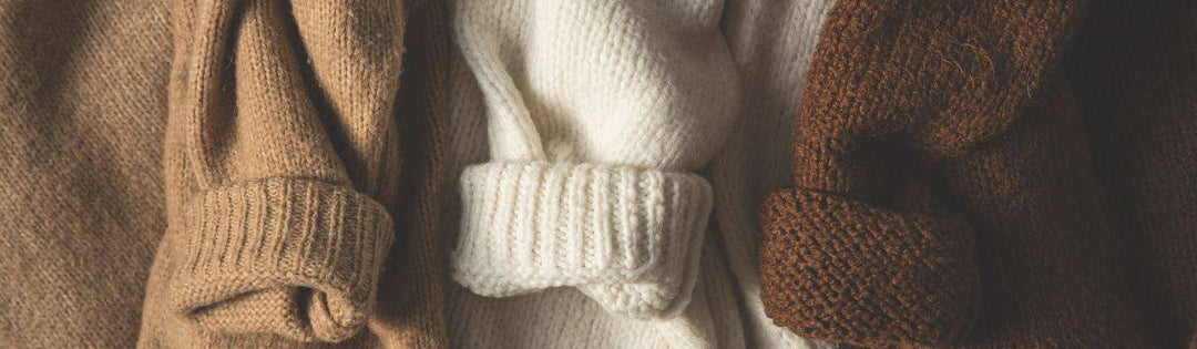 Drei Wollpullover in Beige, Weiß und Dunkelbraun mit hochgekrempelten Ärmeln.